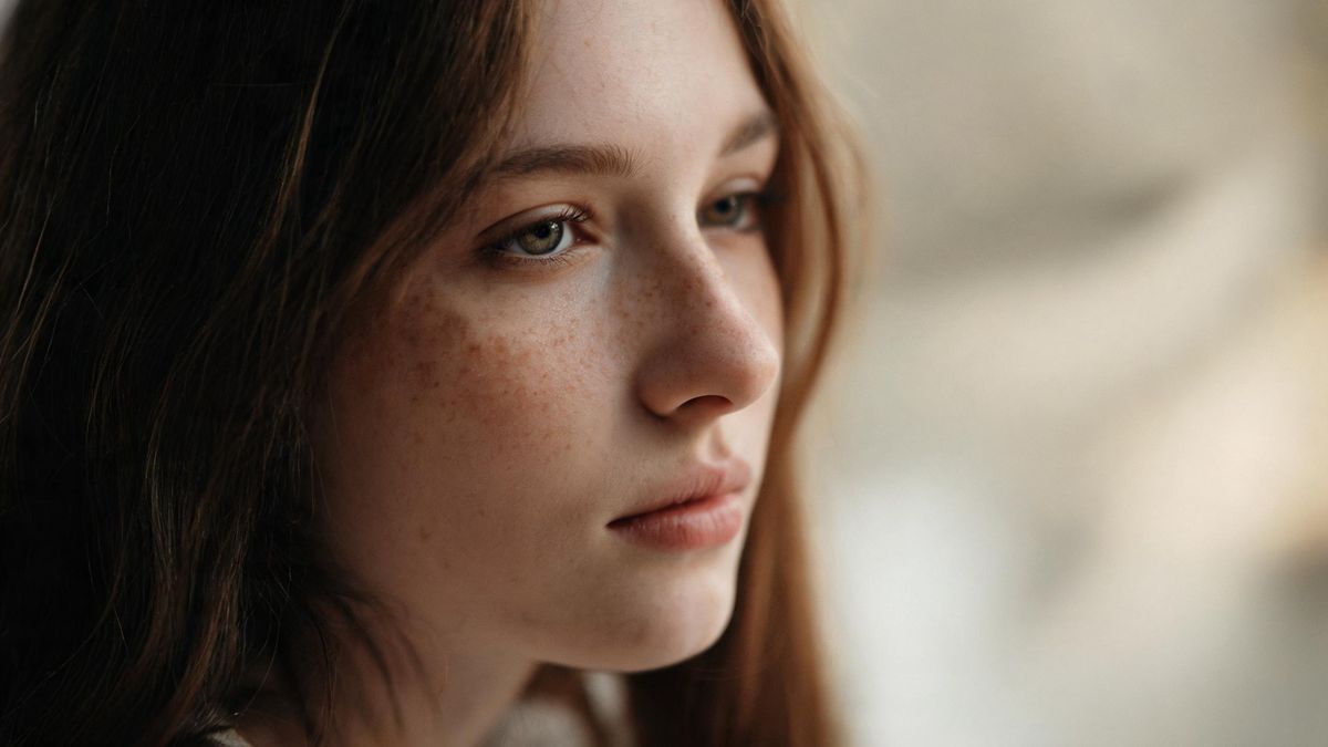 A close view of a face in soft window light with freckles across the nose, the background dissolved into cream