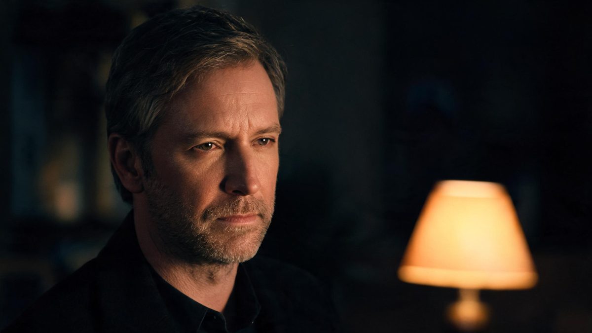 A European man in his forties in a dim room with a single warm lamp catching one side of his face, the rest in soft shadow, no text anywhere