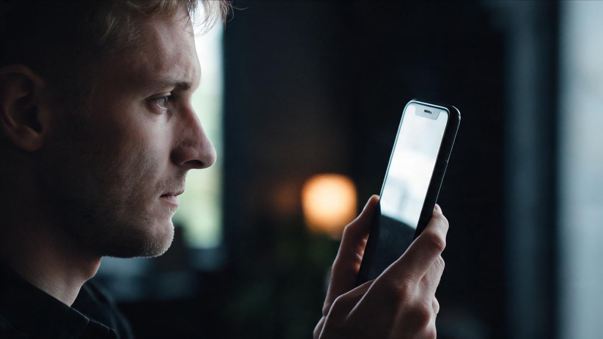 A hand holding a phone at an angle so the screen reflects only a bright window, the device in silhouette, no face visible