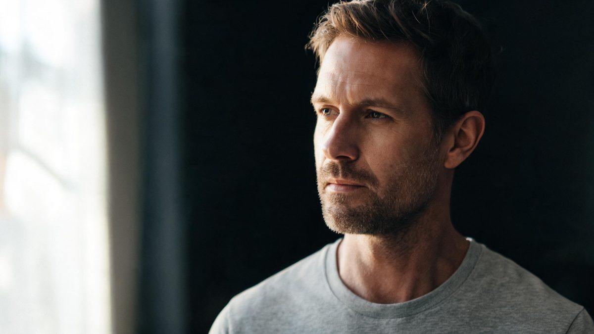 A European man in a plain crew-neck shirt turning his head half towards a soft window, one side of his face in light and the other in gentle shadow, neutral expression, no text anywhere