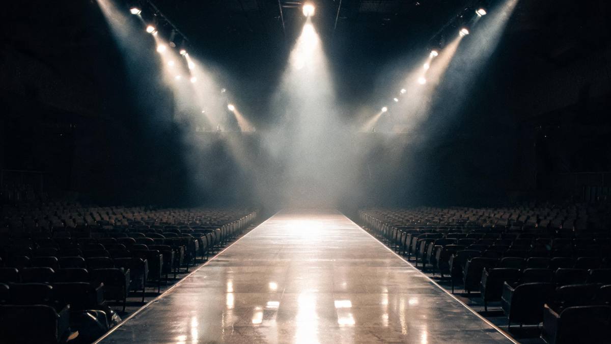 An empty fashion runway with overhead spotlights converging on a polished floor and dark seating on both sides