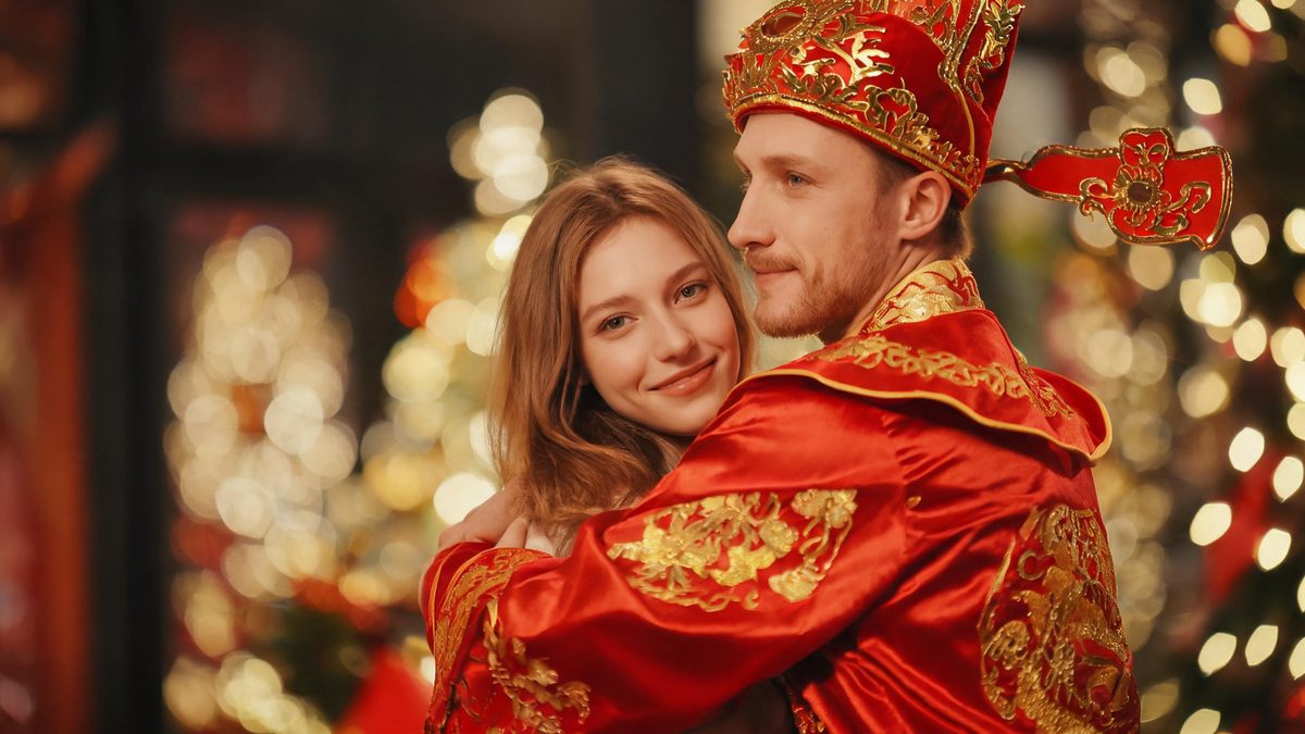 A woman being embraced by a traditional Chinese God of Wealth figure who has just walked into frame
