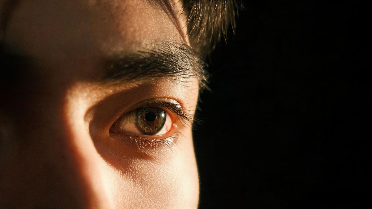 Close-up of a man’s eye and brow in warm lamplight with detail running along the lashes, the background entirely black
