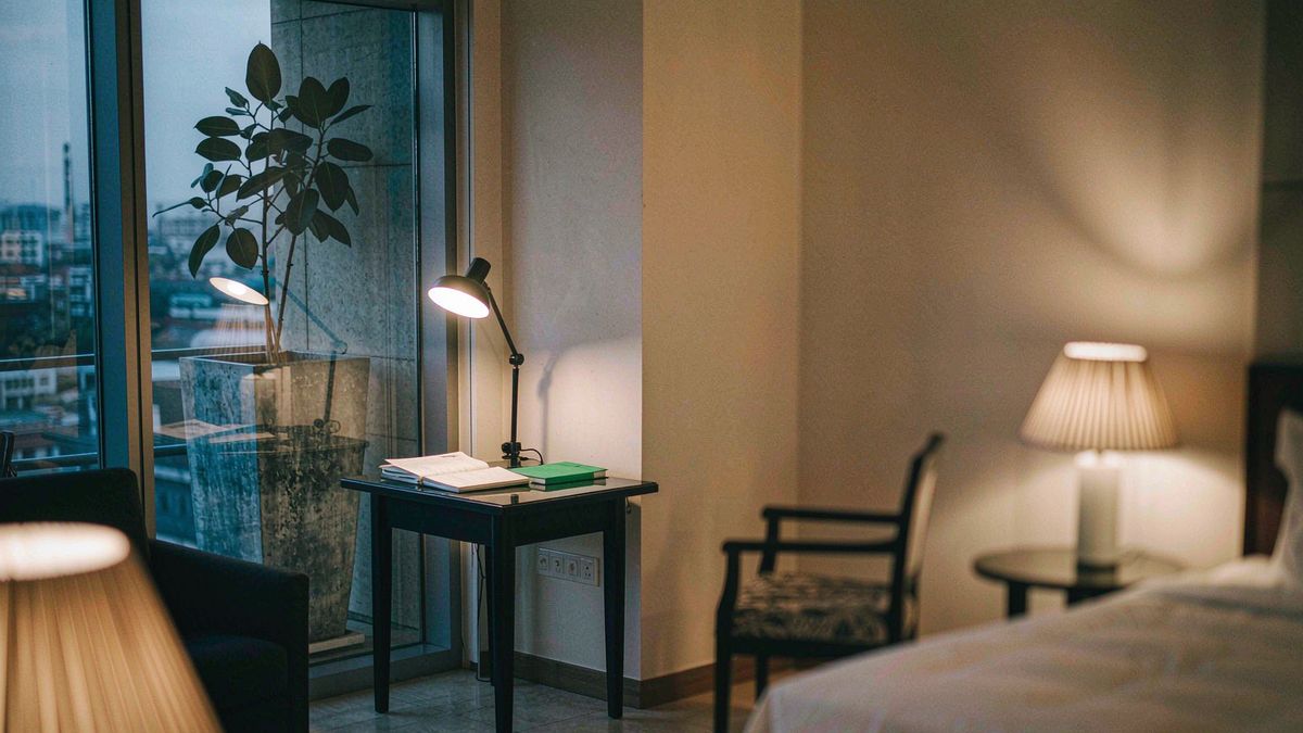 A desk beside a tall window at dusk with a notebook, plant and lamp