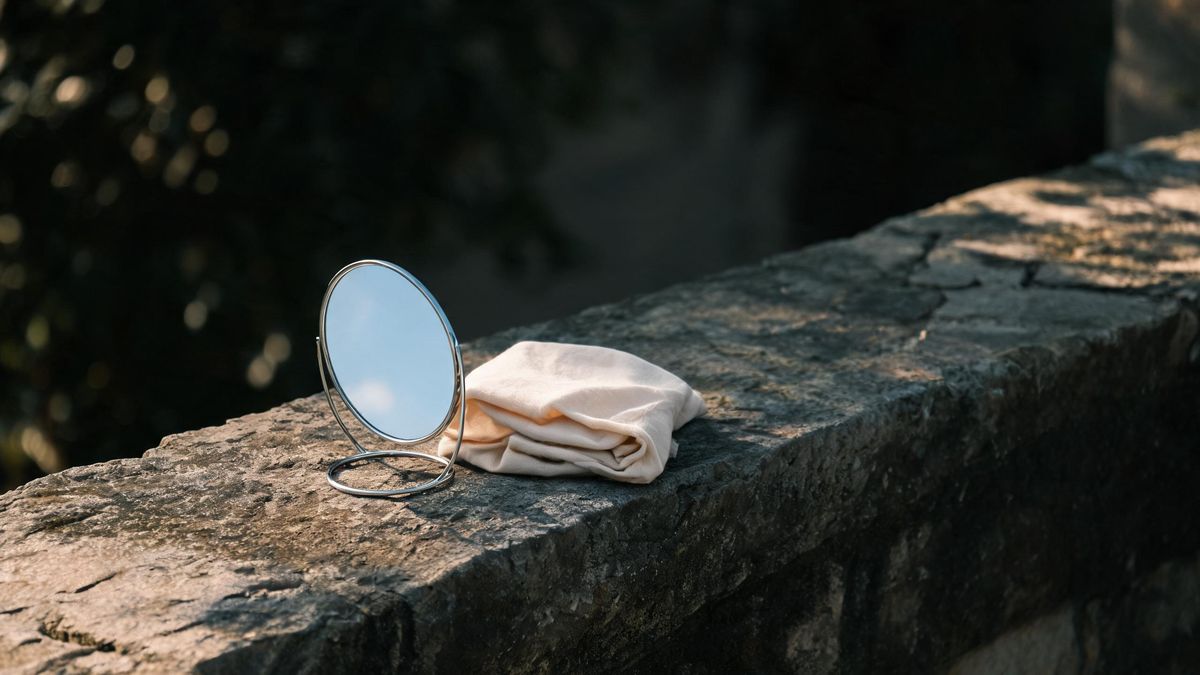 A small round mirror on a stone surface with a folded soft cloth beside it, even daylight, no people