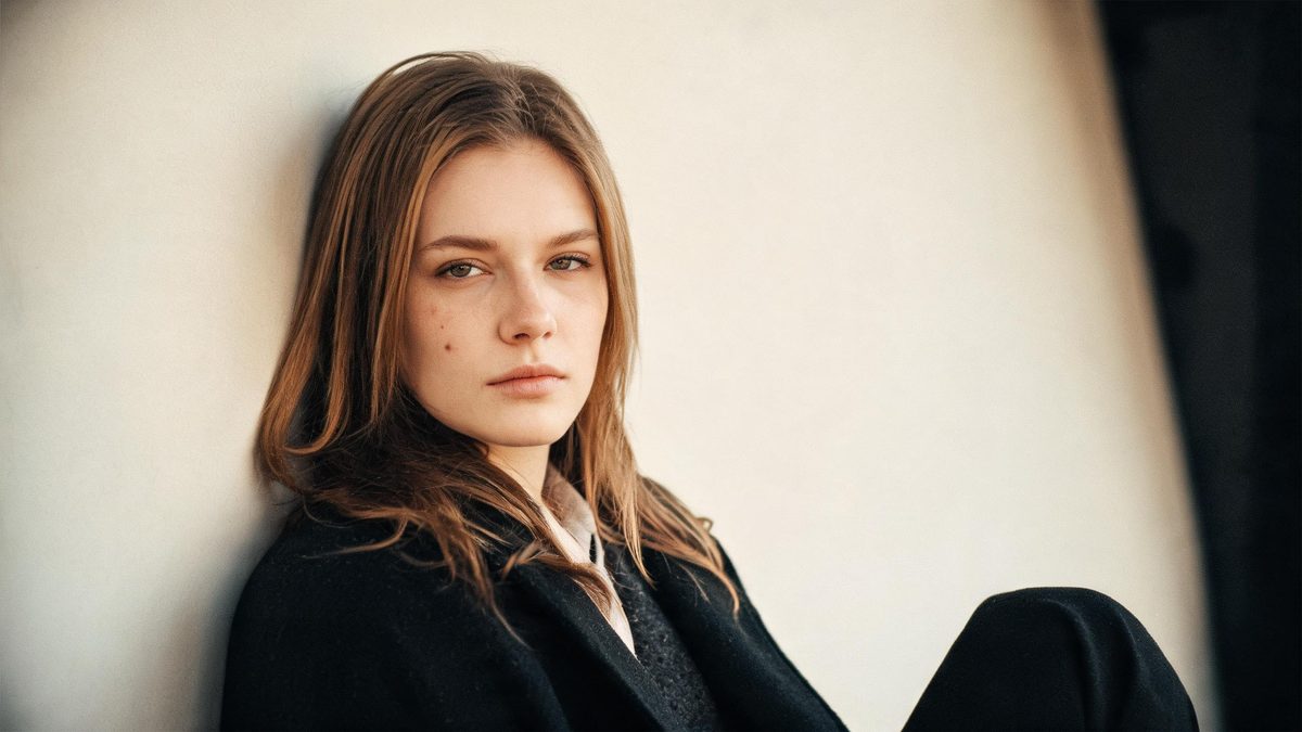 A European woman seated against a pale wall in even daylight, a faint mark on her cheek, calm expression, shallow depth of field, no text anywhere
