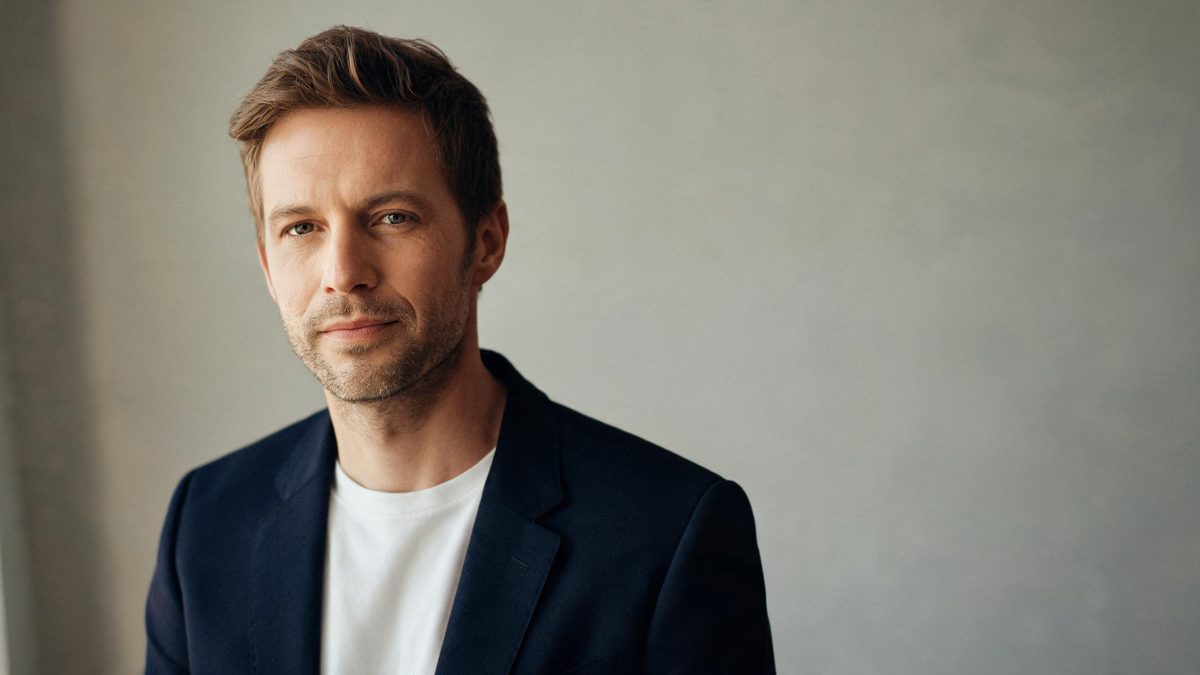 A person in a blazer against a plain neutral wall, even soft light, calm expression.