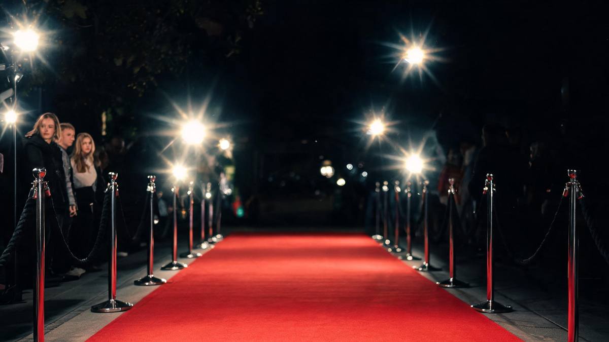 An empty red carpet at night bordered by stanchions and rope with pop-flash bulbs flaring above and deep shadow beyond