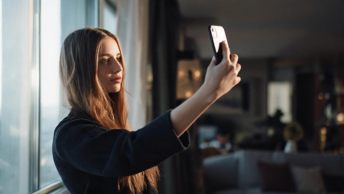 A person holding a phone at arm's length by a window, natural daylight on the face.