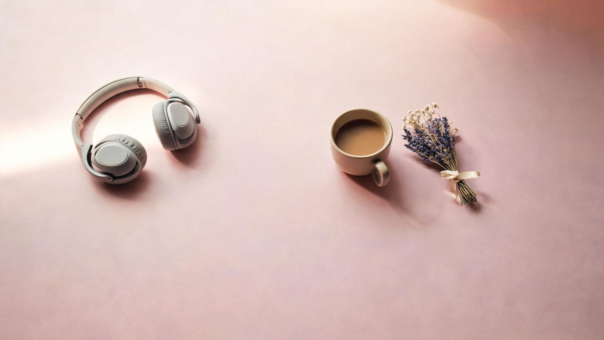 A flat lay of headphones, a coffee cup and dried flowers on a pastel desk.