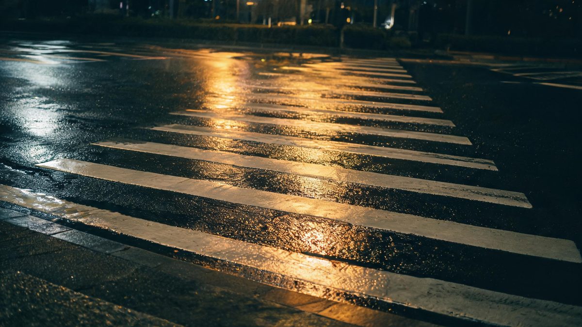 An empty crosswalk at night in rain, wet asphalt reflecting light, no clear faces.