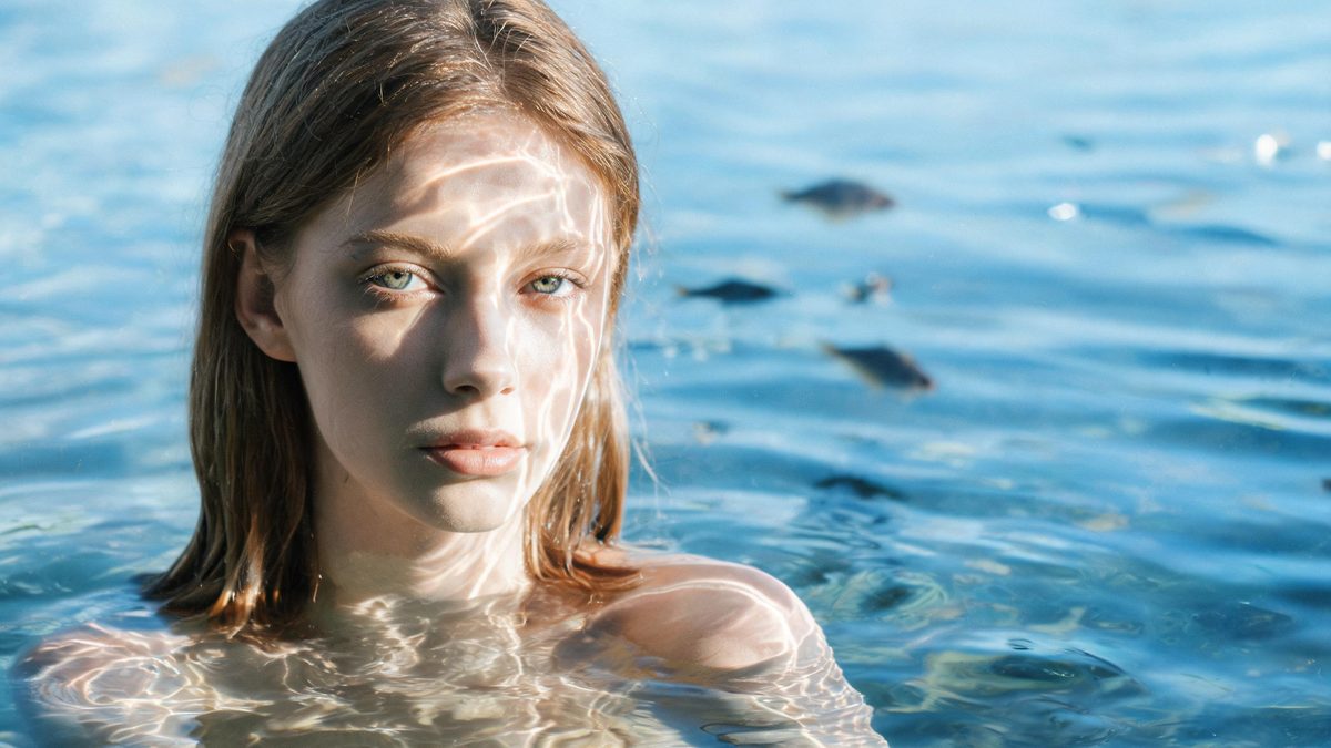 A woman suspended half-body in clear blue water with rippling light patterns across her face and small fish silhouettes behind