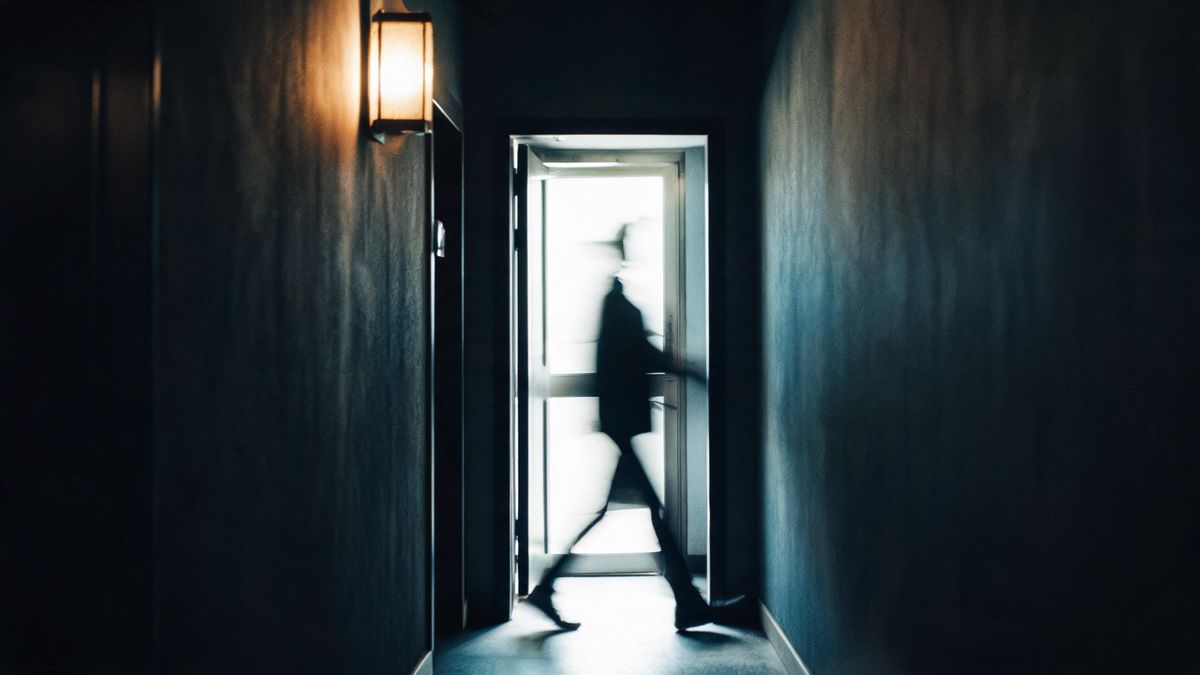 A blurred figure walking through a bright doorway in a dark corridor, the motion rendering as a soft streak