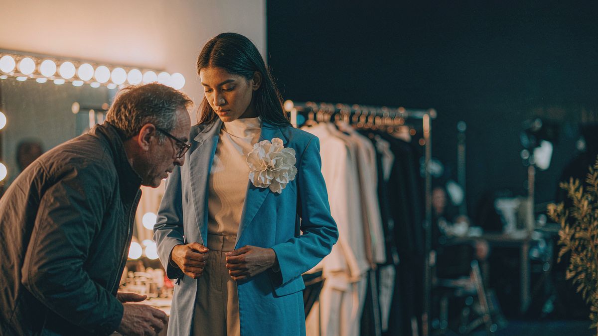 A model in a tailored coat being fitted backstage under mirror lights