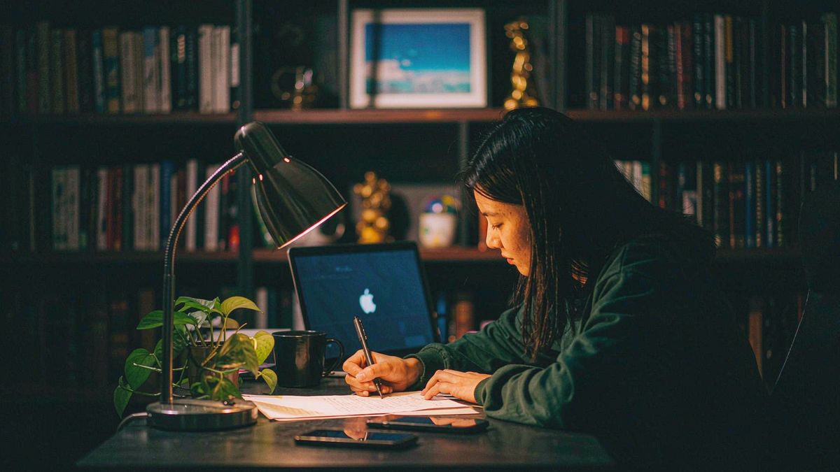 A person writing at a desk beside a laptop in a lamp-lit study at night