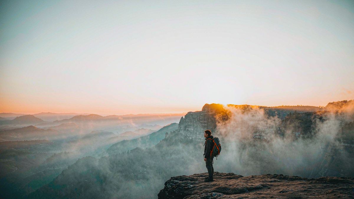 A traveller with a backpack on a ledge above a misty mountain valley at dawn