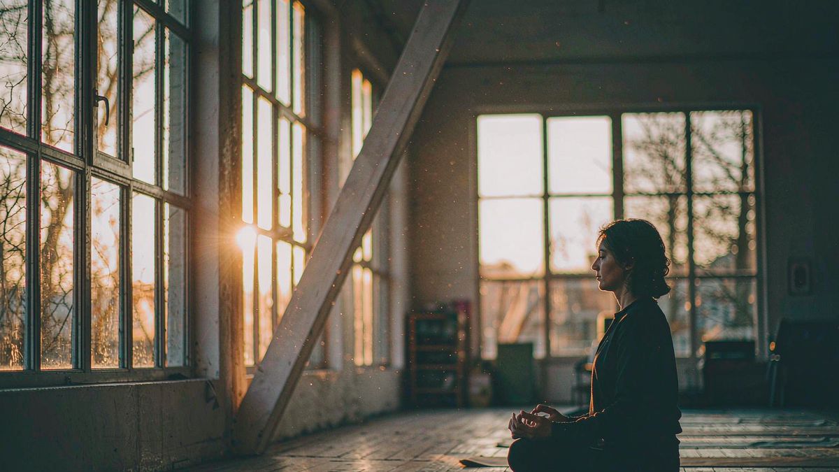 A person seated on a yoga mat in sunrise light streaming through tall windows