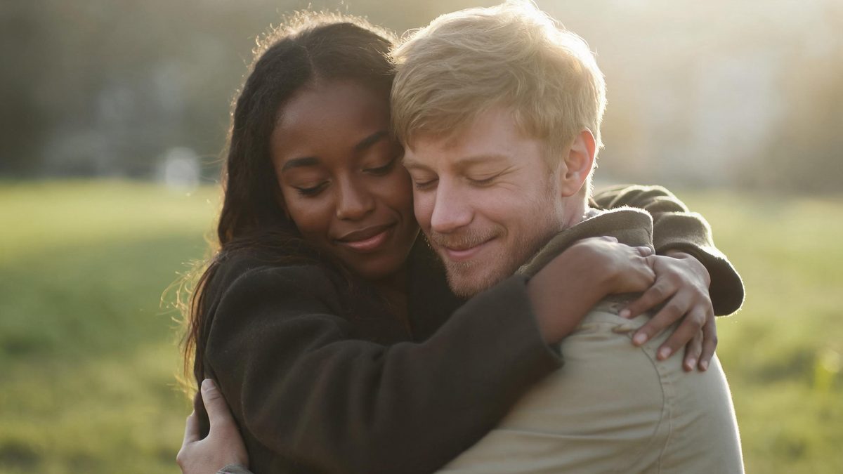 Two friends in a warm embrace outdoors, backlit by late afternoon sun against an empty meadow