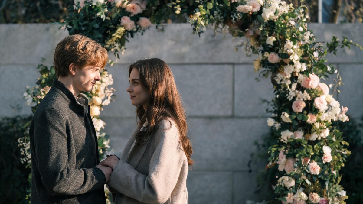 Two people under a flower arch in soft daylight, a neutral stone wall behind.