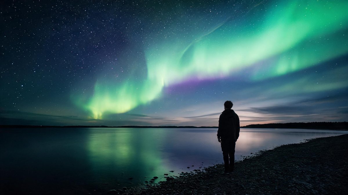 Green and violet aurora light over a dark lake shore with a lone figure standing at the water edge