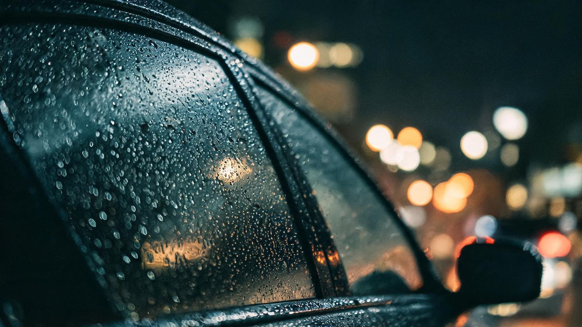 Close-up of a rain-covered windscreen with city lights behind it, sharp droplets against a soft glow