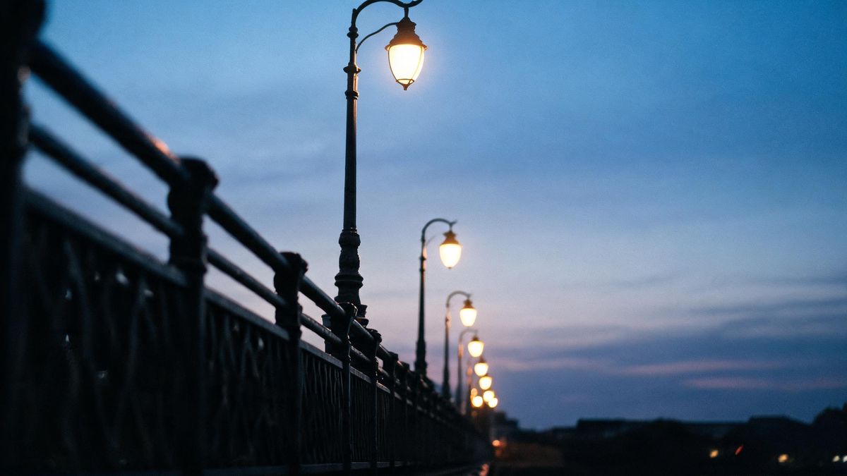 A bridge railing at dusk with lamp posts receding into the distance, warm light against a cooling sky