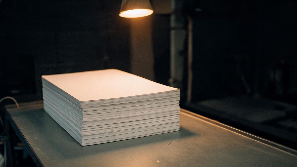 A tall stack of photographic paper on a metal table in a dim print room, warm light from a single overhead fixture