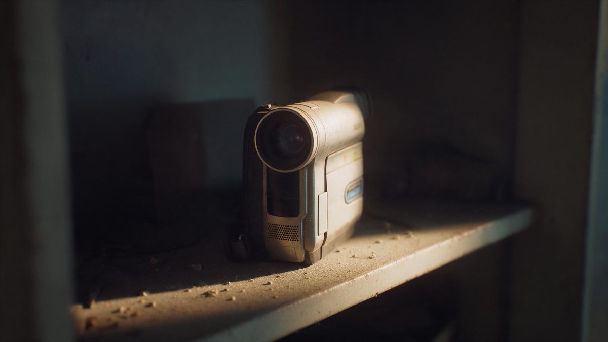 An old camcorder on a dusty shelf in a dim room, warm light grazing its casing, the rest of the room in darkness