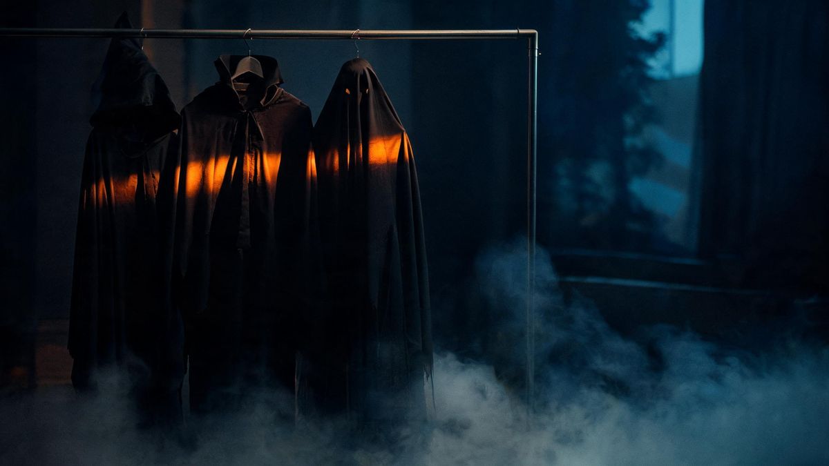 Three dark costumes hanging on a rail in a dim room, caught by one band of warm light