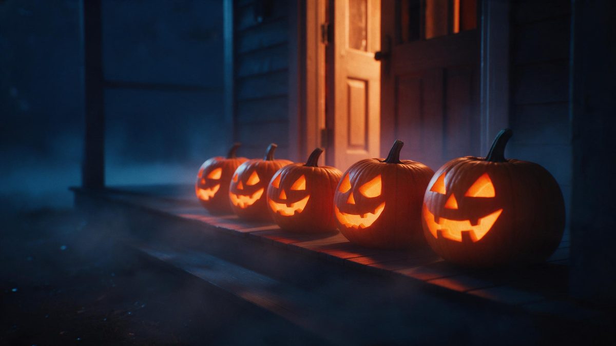 Five carved pumpkins glowing in a row along a wooden porch at night, a door half open behind them with light spilling out