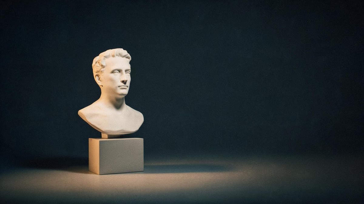 A matte white presenter bust on a low pedestal in an empty studio, lit by one warm key light against a charcoal backdrop