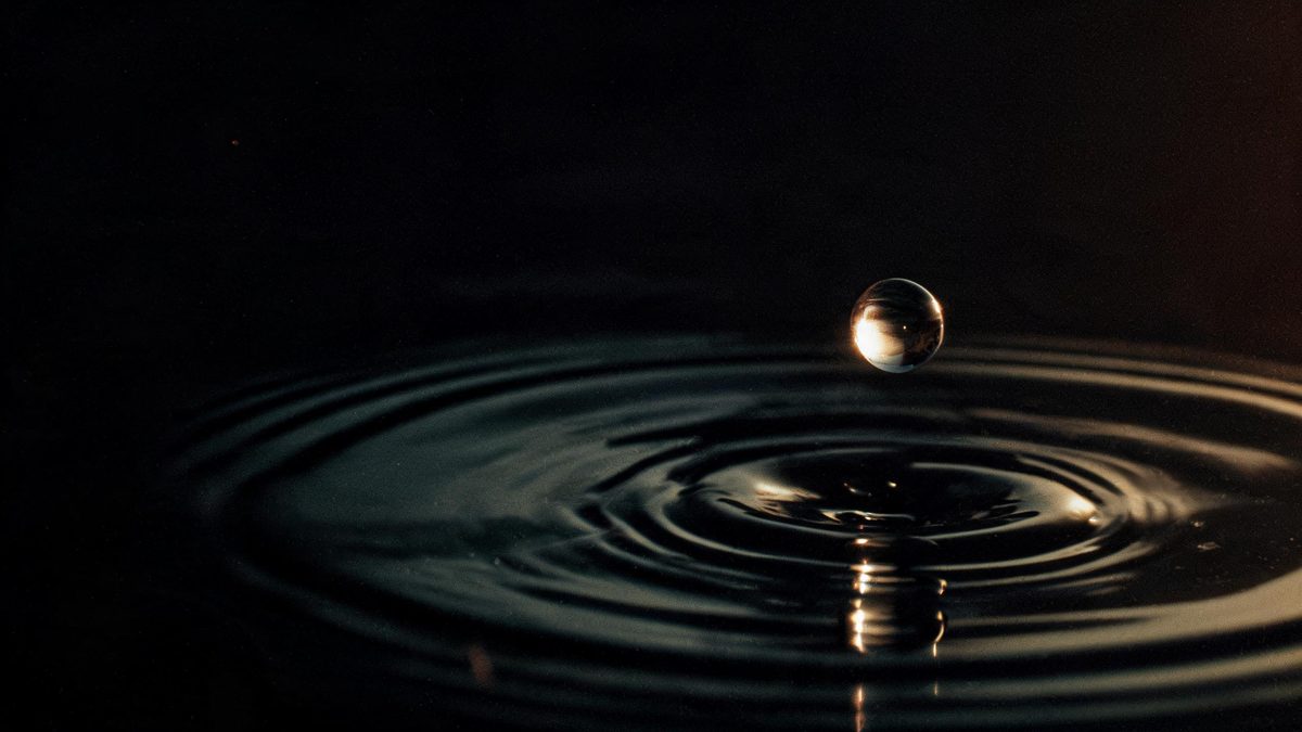 A single water droplet frozen above still dark water with concentric ripples and a hard side light