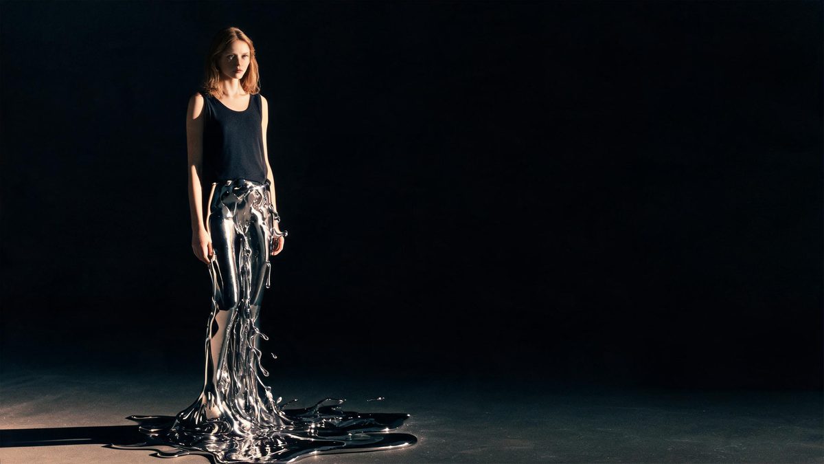 A figure whose lower body dissolves into flowing liquid chrome pooling on a dark floor under dramatic side light