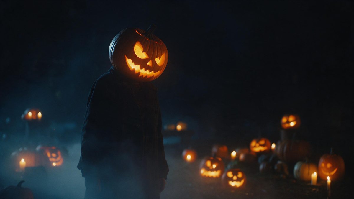 A figure in a dark coat whose head is a glowing carved pumpkin, set against black