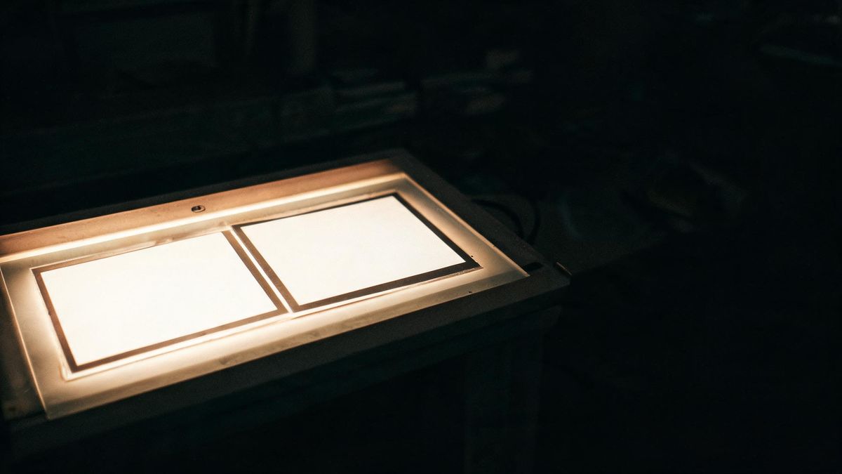 A light table in a darkroom with two prints laid on the glass, their edges glowing while the room stays dark