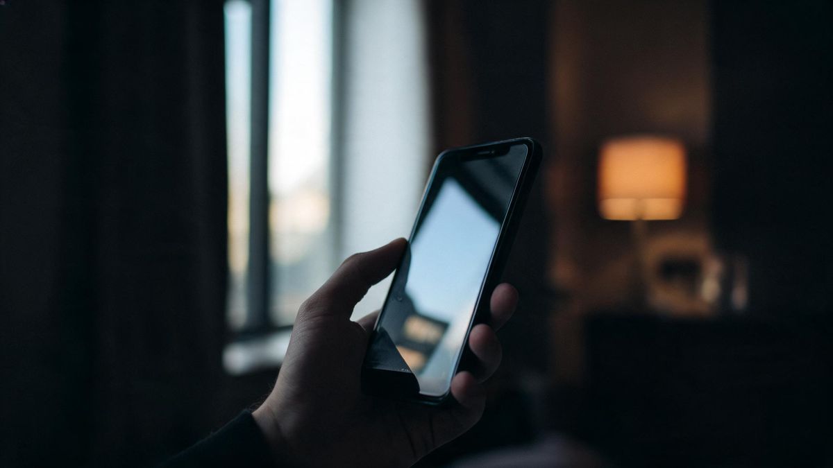 A hand holding a phone at an angle so the surface reflects only a window, the device itself in silhouette