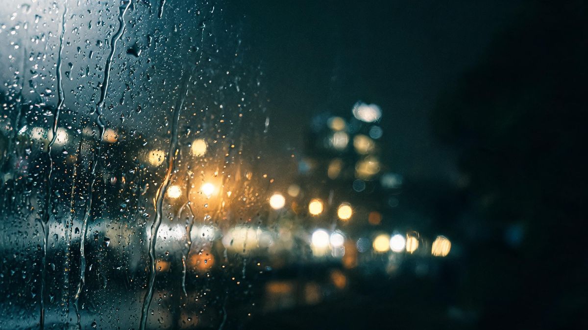 Rain on a window pane with a blurred city beyond, droplets sharp in the foreground and soft light behind