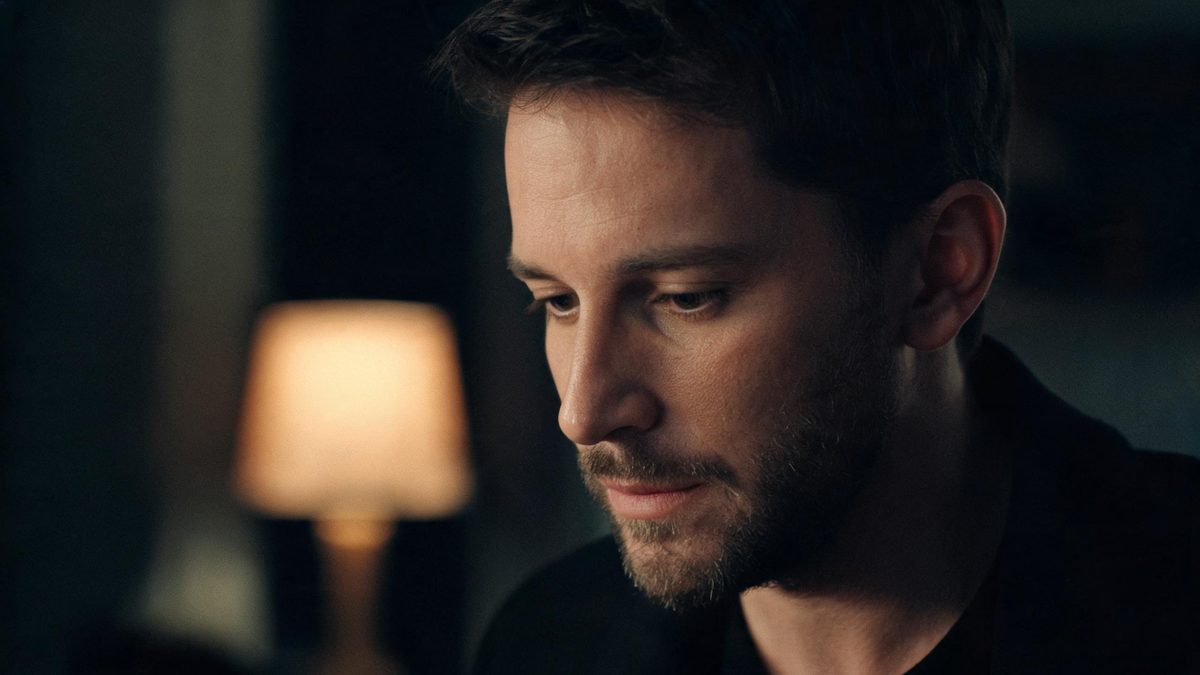 Close-up of a man’s face at a three-quarter angle lit by a single warm lamp, eyes downcast
