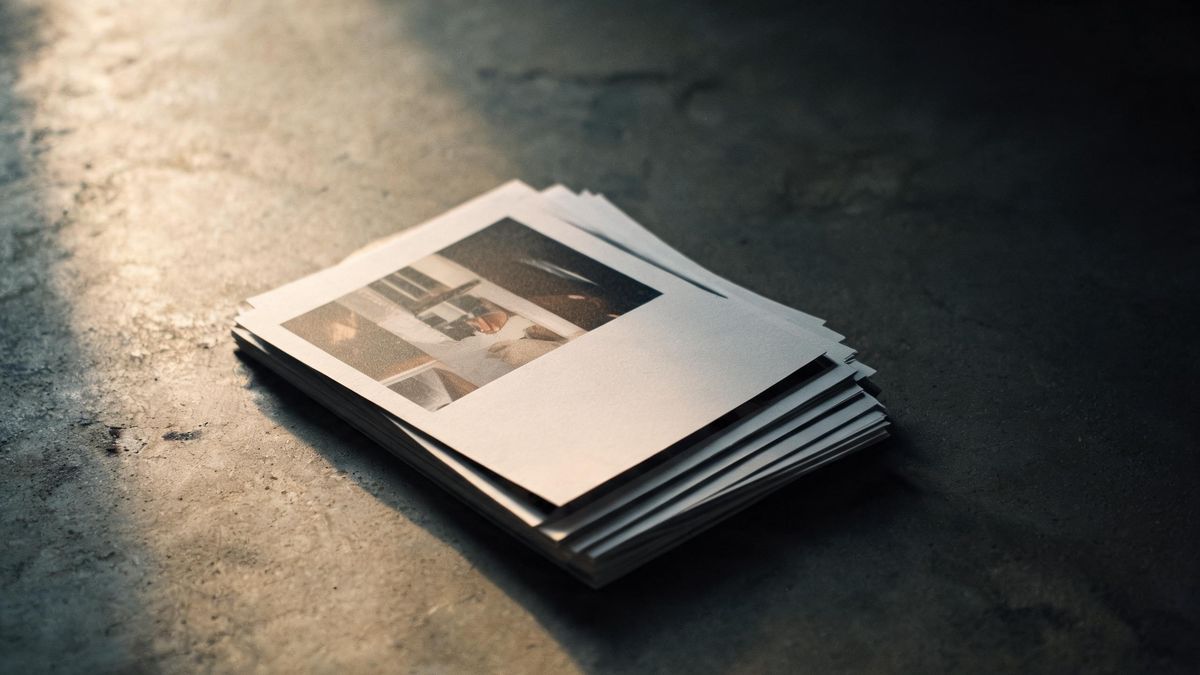 A stack of photographic prints on a concrete floor seen from above, the top print lit from one side and the edges fading into shadow