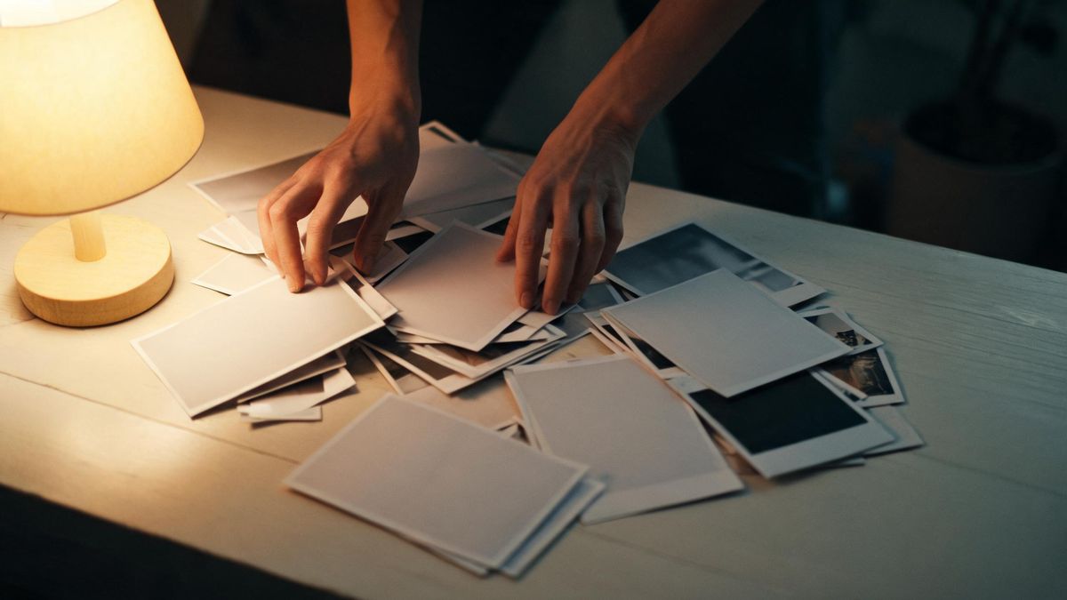 Two hands sorting loose photographic prints on a pale table under a warm lamp set to the left